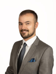 Uśmiechnięty, krótko strzyżony mężczyzna z zarostem w szarym garniturze.
