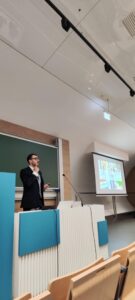 Sala konwersacji, na tle tablicy mężczyzna przemawiający i prezentujący zagadnienia tematyczne