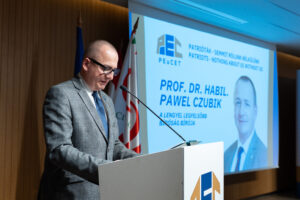 Prof. Czubik przemawia przy mównicy, w tle widać ekran z prezentacją