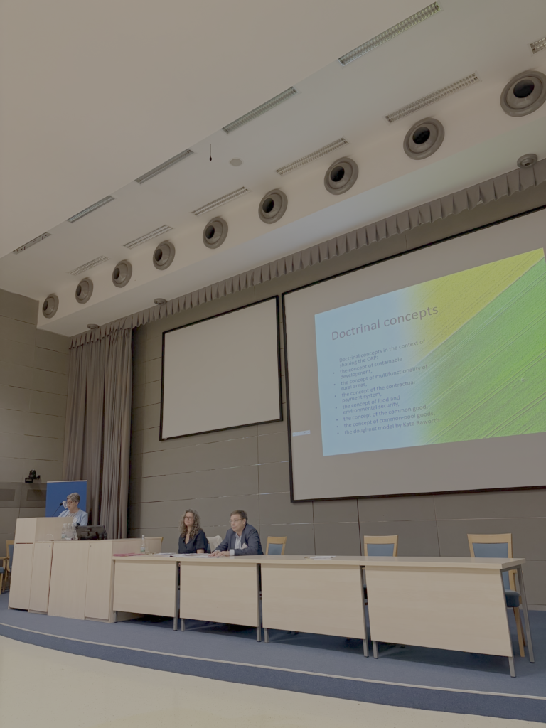 sala konferencyjna, duża tablica multimedialna z wyświetloną prezentacją, długi stół konferencyjny, dwie osoby przy stole, kobieta stoi przy mównicy