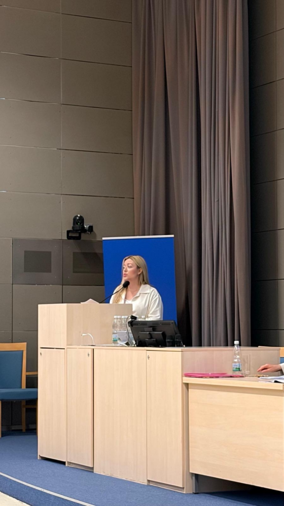 mównica, kobieta w jasnej marynarce, w tle kurtyna i niebieska tablica